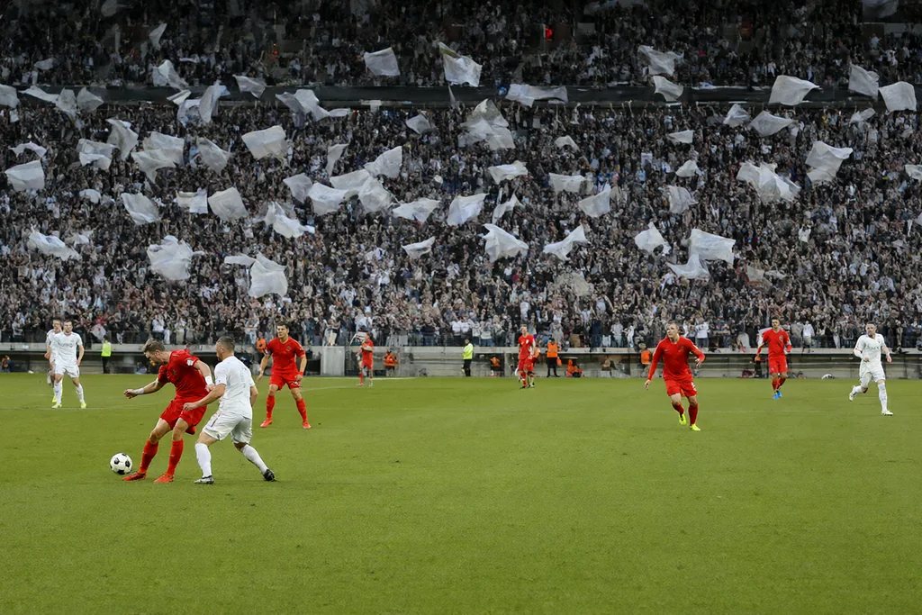 Fans hinter dem Tor mit wehenden Fahnen auf der vollen Tribüne im Flutlicht